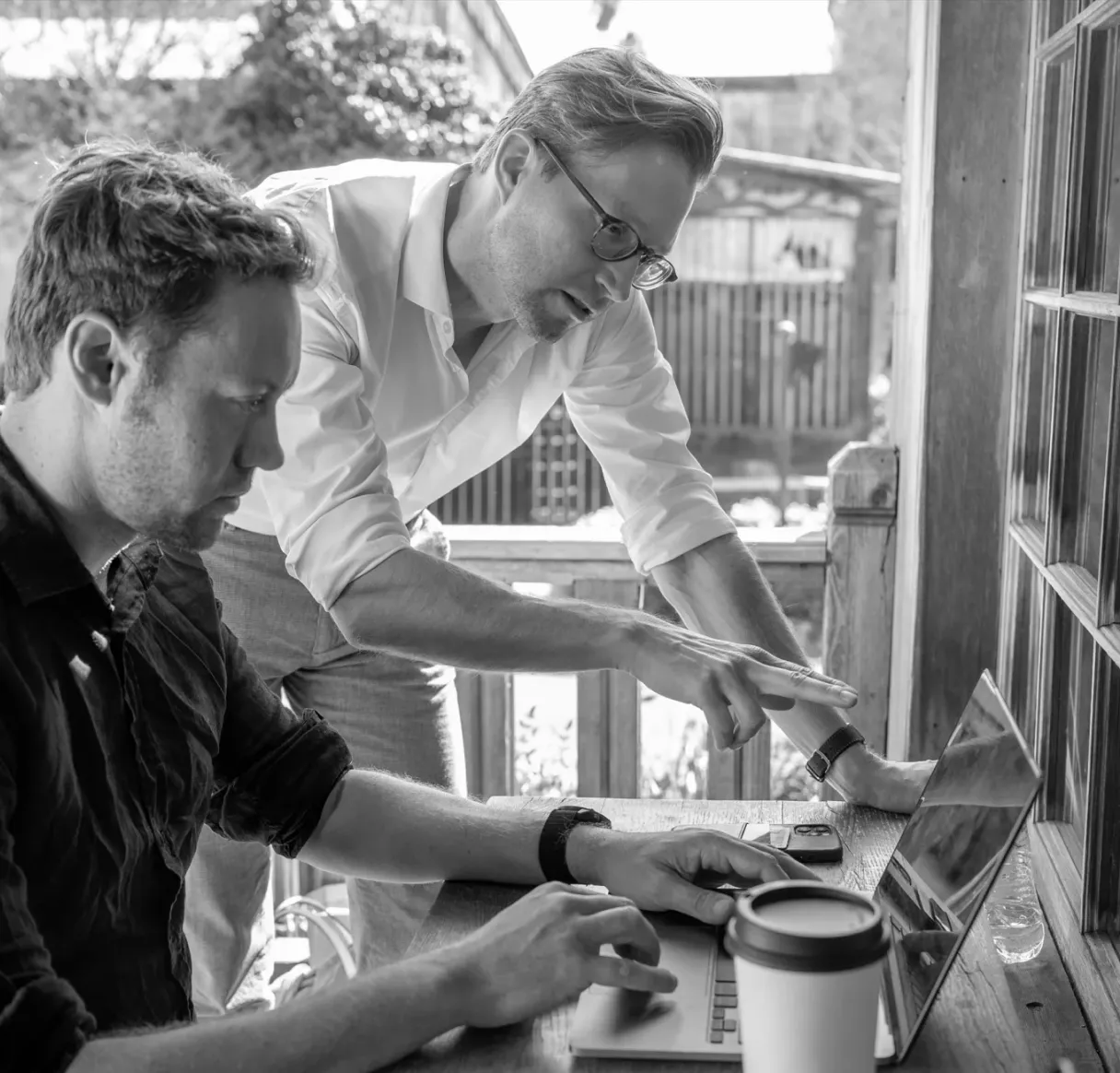 A black and white photo capturing a focused coaching moment. One man stands and points at a laptop screen to guide his seated colleague, illustrating personalized mentorship, teamwork, and a detailed strategy review.