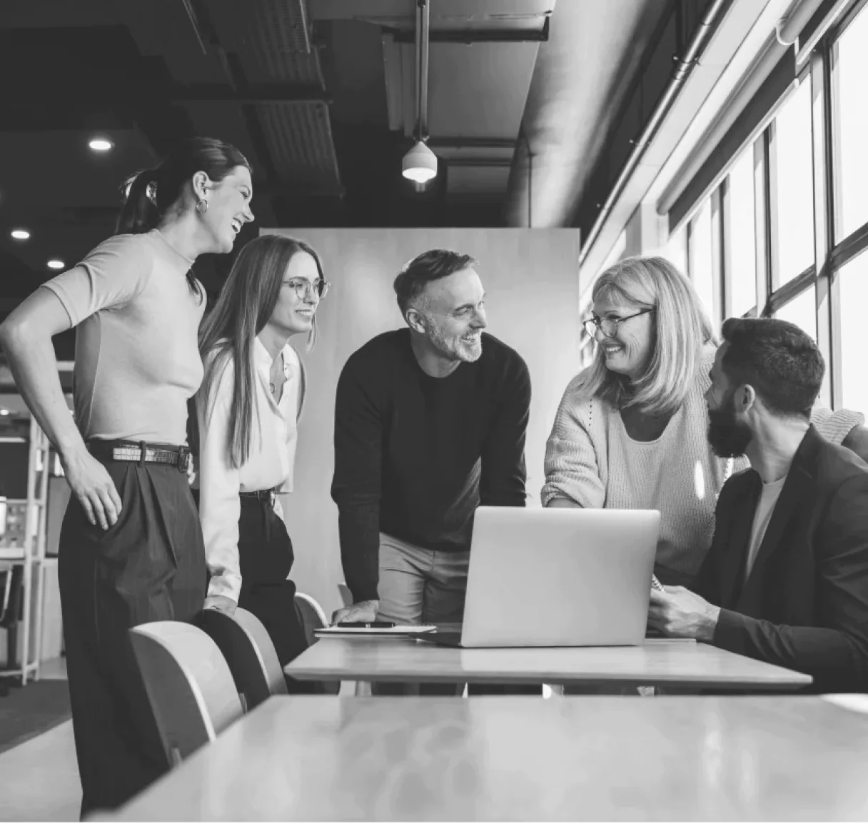 A black and white photo showing a group of five smiling colleagues gathered around a table and looking at a laptop. The image captures a moment of successful collaboration and positive team chemistry in a modern office environment.
