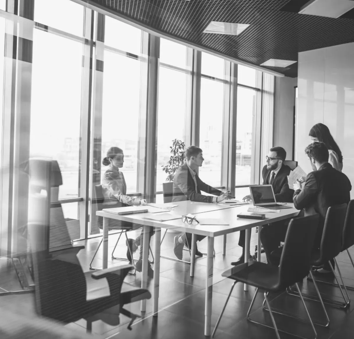 A black and white photo looking into a glass conference room where a team of five professionals is engaged in a focused discussion. They are gathered around a table with laptops and papers, with large city-view windows behind them.