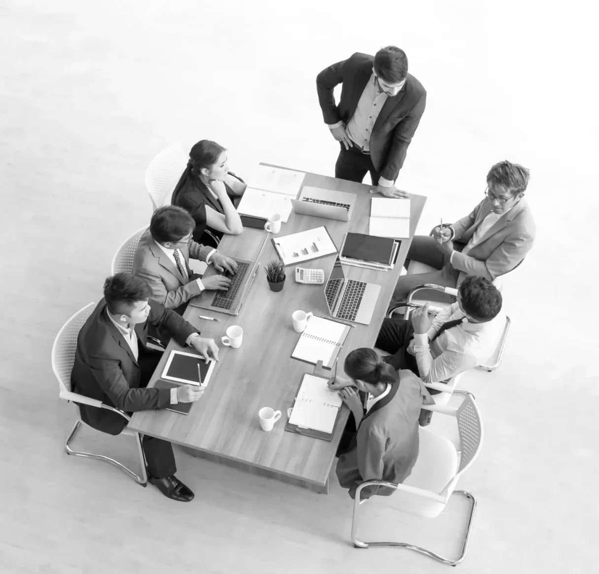 A black and white overhead shot of a formal business meeting. One person stands at the head of a long table, addressing six seated colleagues who are working with laptops, tablets, and documents in a focused strategy session.