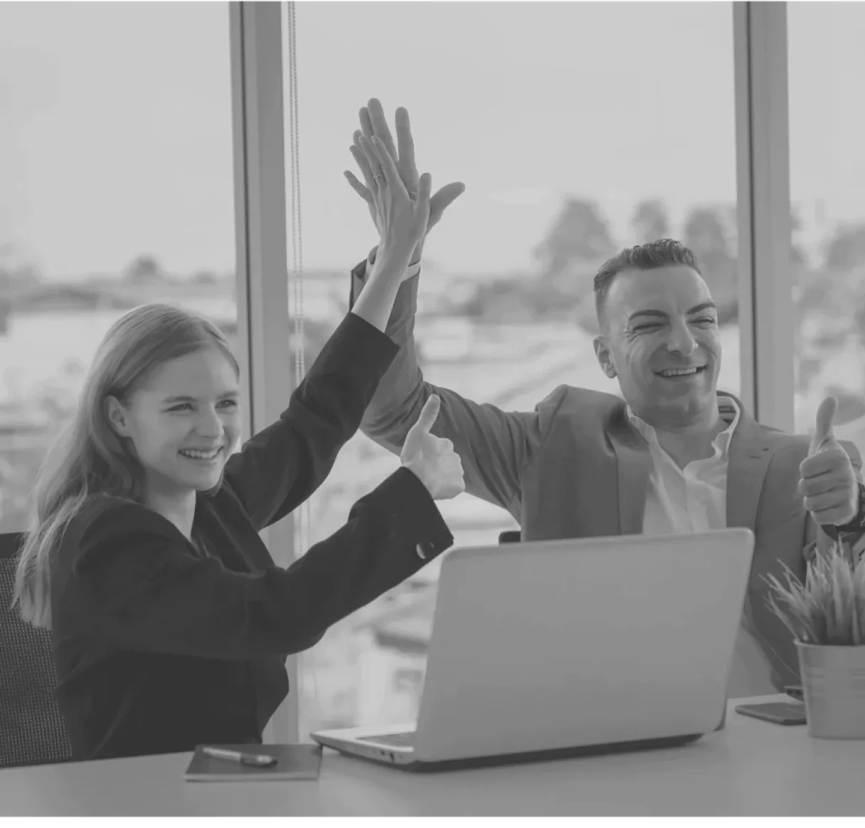 A black and white photo of a man and a woman at an office desk, celebrating a business success. They are smiling broadly and giving each other a high-five over their laptop, symbolizing successful teamwork and goal attainment.