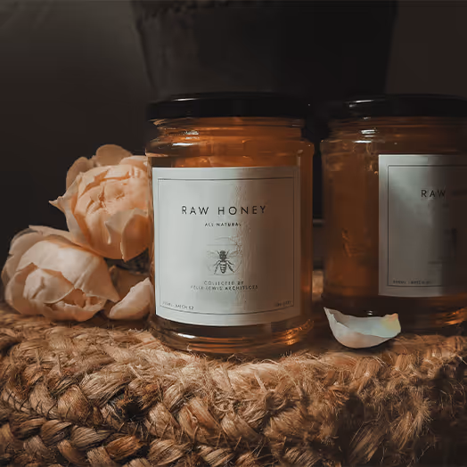 A product shot of FLA Raw Honey. It shows a couple of jars of honey positioned next to some flowers