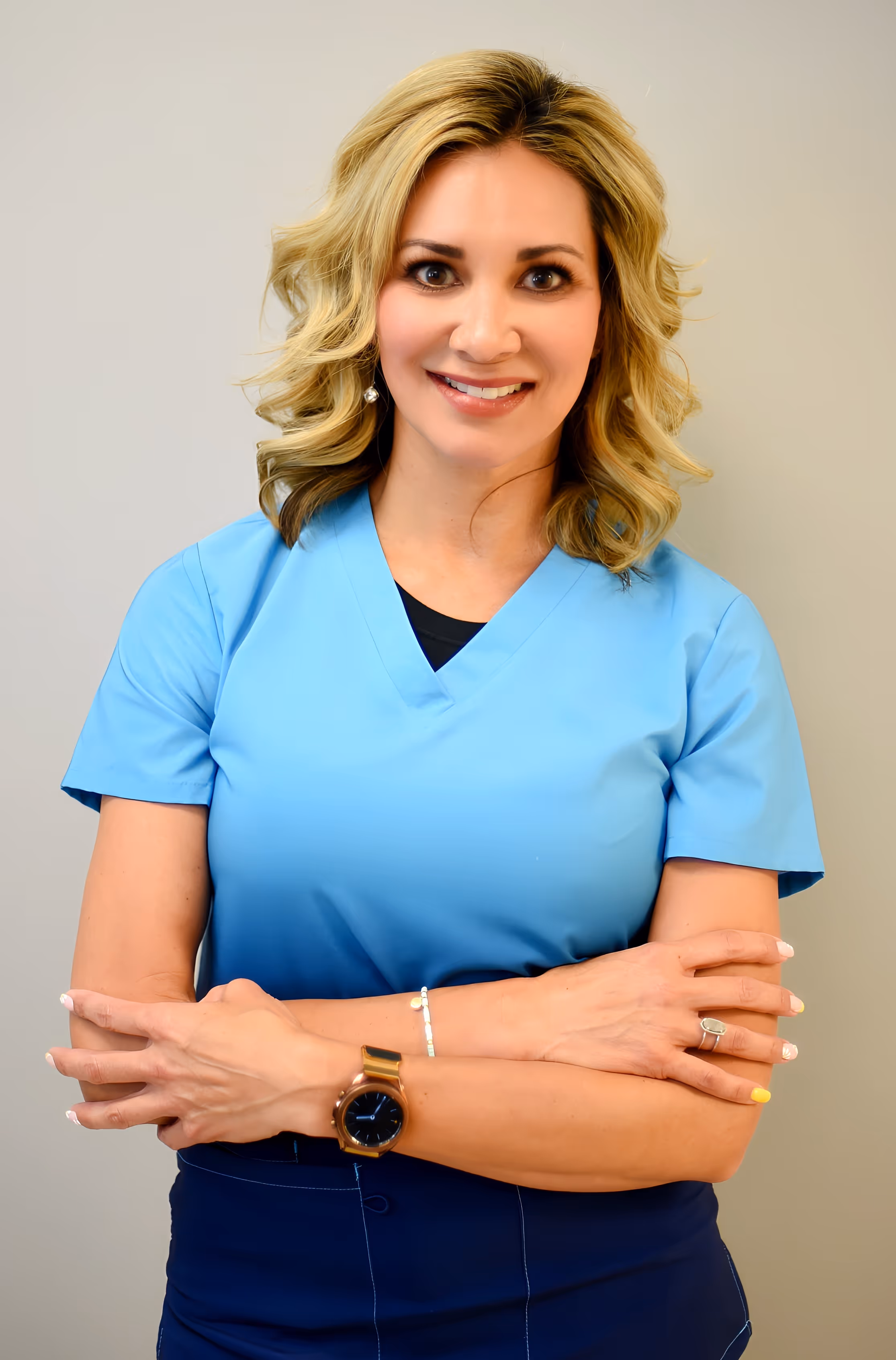 Smiling woman with blonde curly hair wearing blue medical scrubs and a smartwatch, standing with arms crossed.