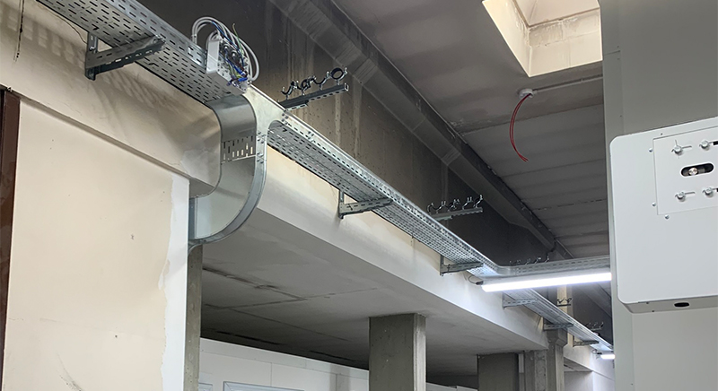 Industrial ceiling with metal cable trays holding electrical wiring and fluorescent lighting fixtures in a building under construction.