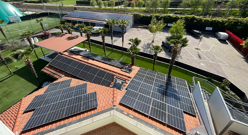 Aerial view of solar panels installed on terracotta tiled roofs surrounded by palm trees, with a parking lot and sports courts in the background.