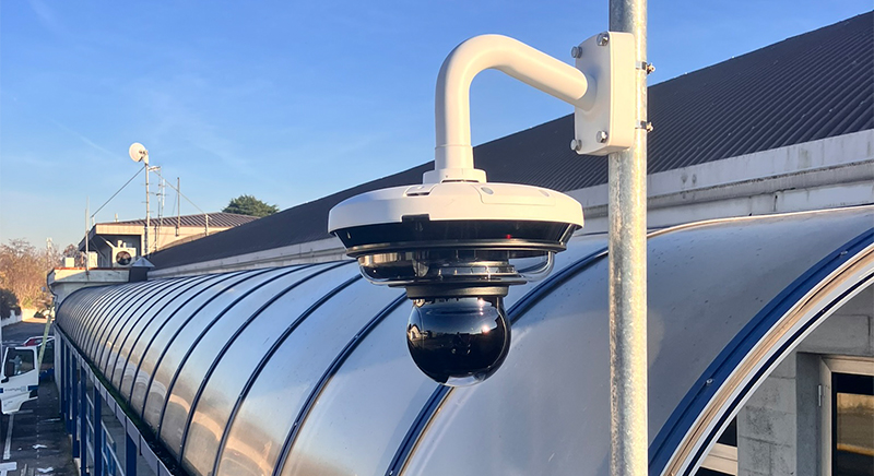 Close-up of a white and black security camera mounted on a pole with a curved metal roof and blue sky in the background.