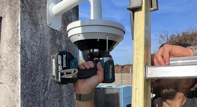 Person installing a white security camera on a wall using a Makita cordless drill with a ladder nearby.