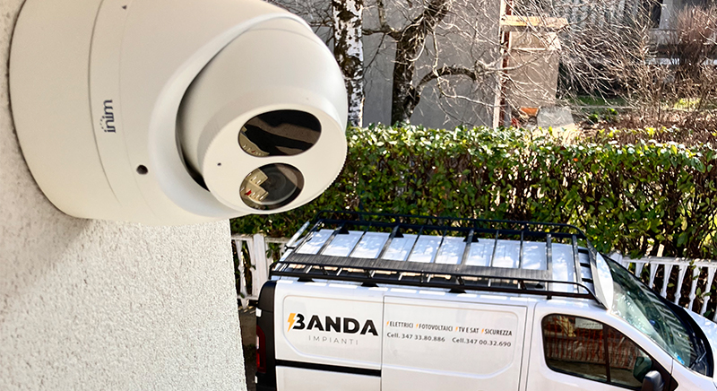 White security camera mounted on a wall overlooking a white van with a roof rack parked near a green hedge and buildings.