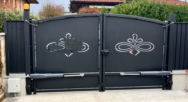 Modern black metal double gate with decorative symmetrical cutout designs and automated opening mechanisms at the bottom.