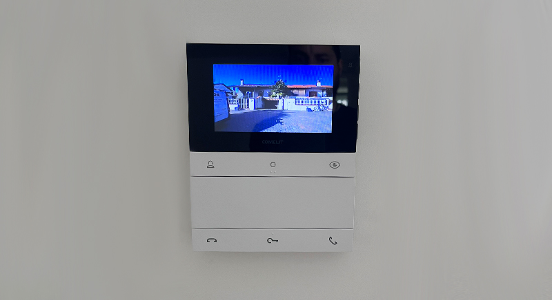 Wall-mounted video intercom showing an outdoor view of a driveway and house entrance.