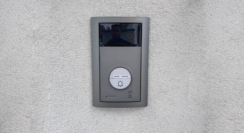 Modern gray video doorbell with a small screen and bell button mounted on a textured light gray wall.