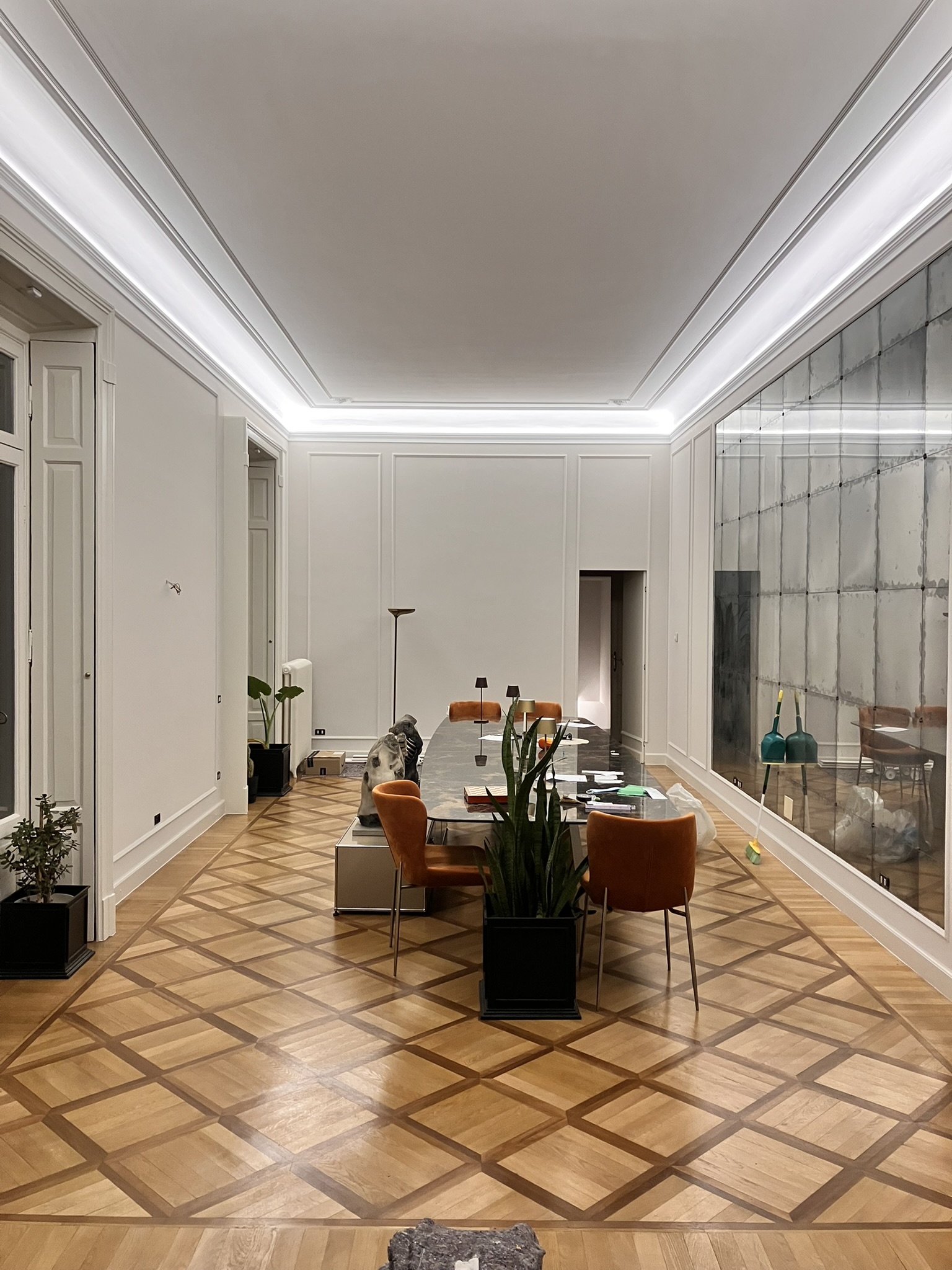 A modern dining room with parquet wood flooring, a long marble table surrounded by orange chairs, plants, and a large reflective wall panel.
