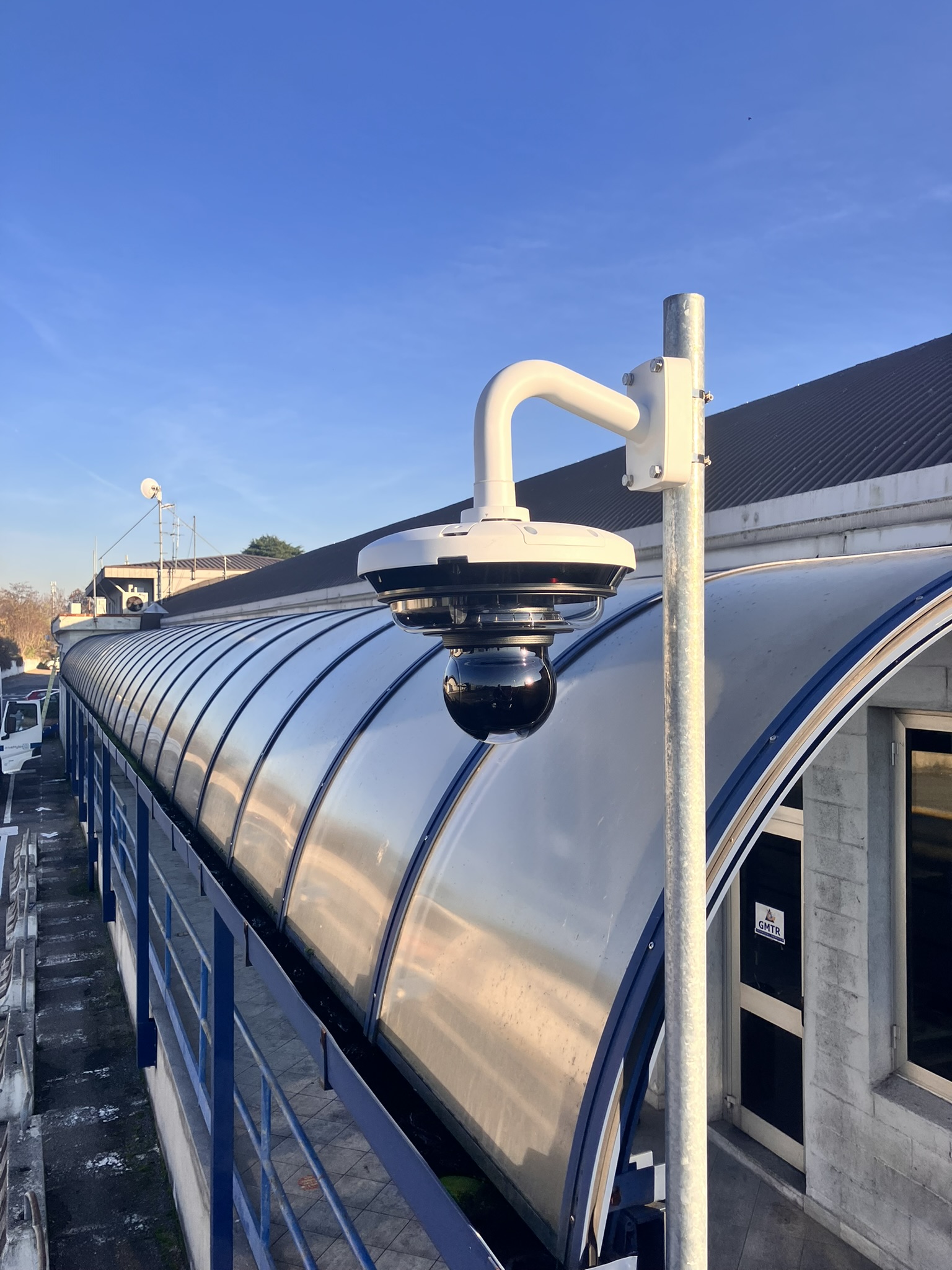 White security dome camera mounted on a pole outside a building with a corrugated metal roof under a clear blue sky.