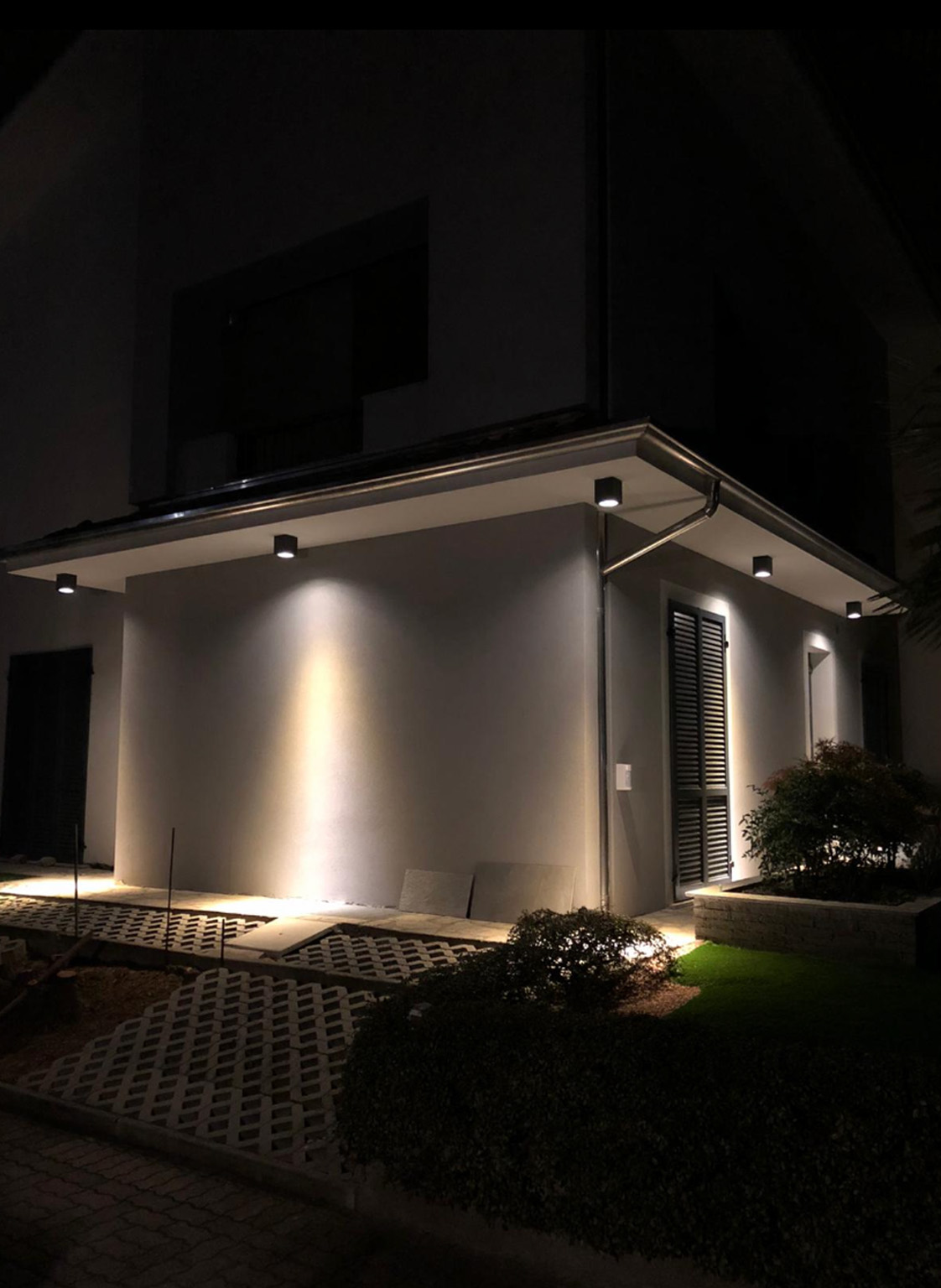 A modern house exterior at night illuminated by multiple wall-mounted lights highlighting the walls, pathway, and shrubs.