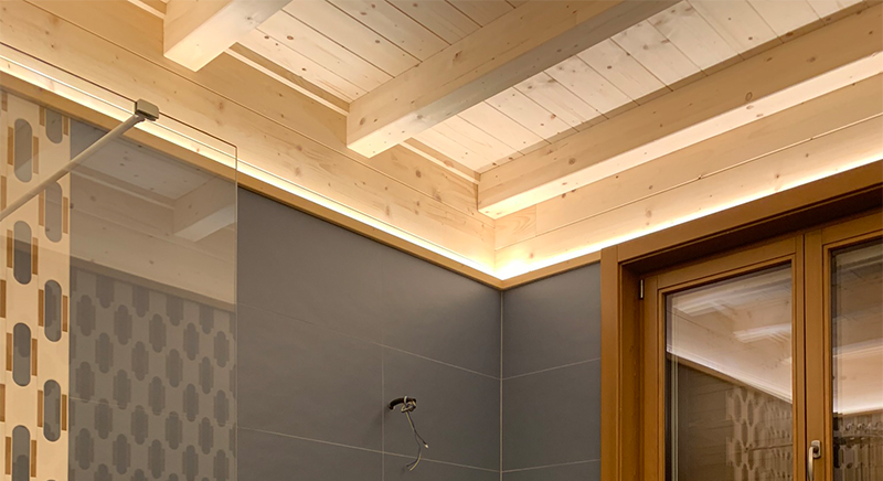 Modern interior corner with wooden ceiling beams, LED strip lighting, dark tiled wall, and glass door.