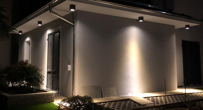 Modern house corner at night illuminated by wall-mounted exterior lights with shadows cast on a white wall and landscaping including bushes and tiled ground.