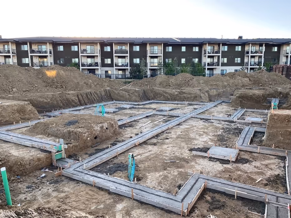 Construction site with concrete foundation footings laid out and dirt piles surrounding it, apartment building in the background.