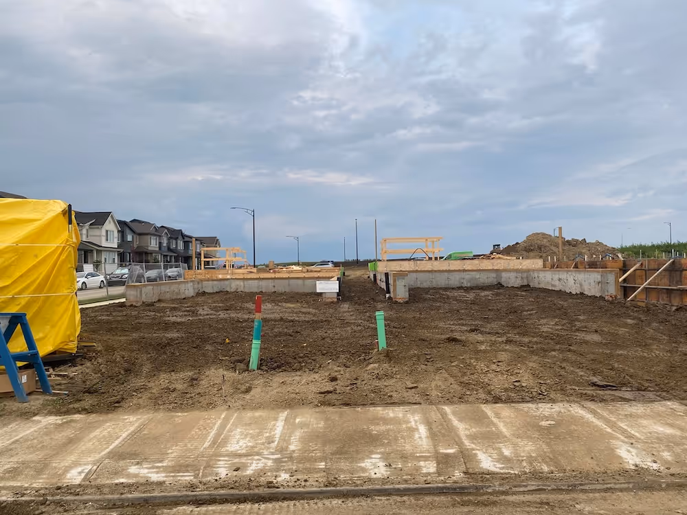 Cleared construction site with concrete foundation walls and exposed dirt, surrounded by suburban houses under a cloudy sky.