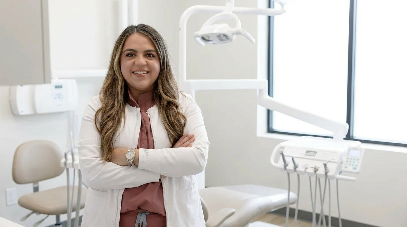 Dr. Alex Franco, in clinical attire, stands confidently in a bright, modern dental office, smiling directly at the camera with her arms crossed.