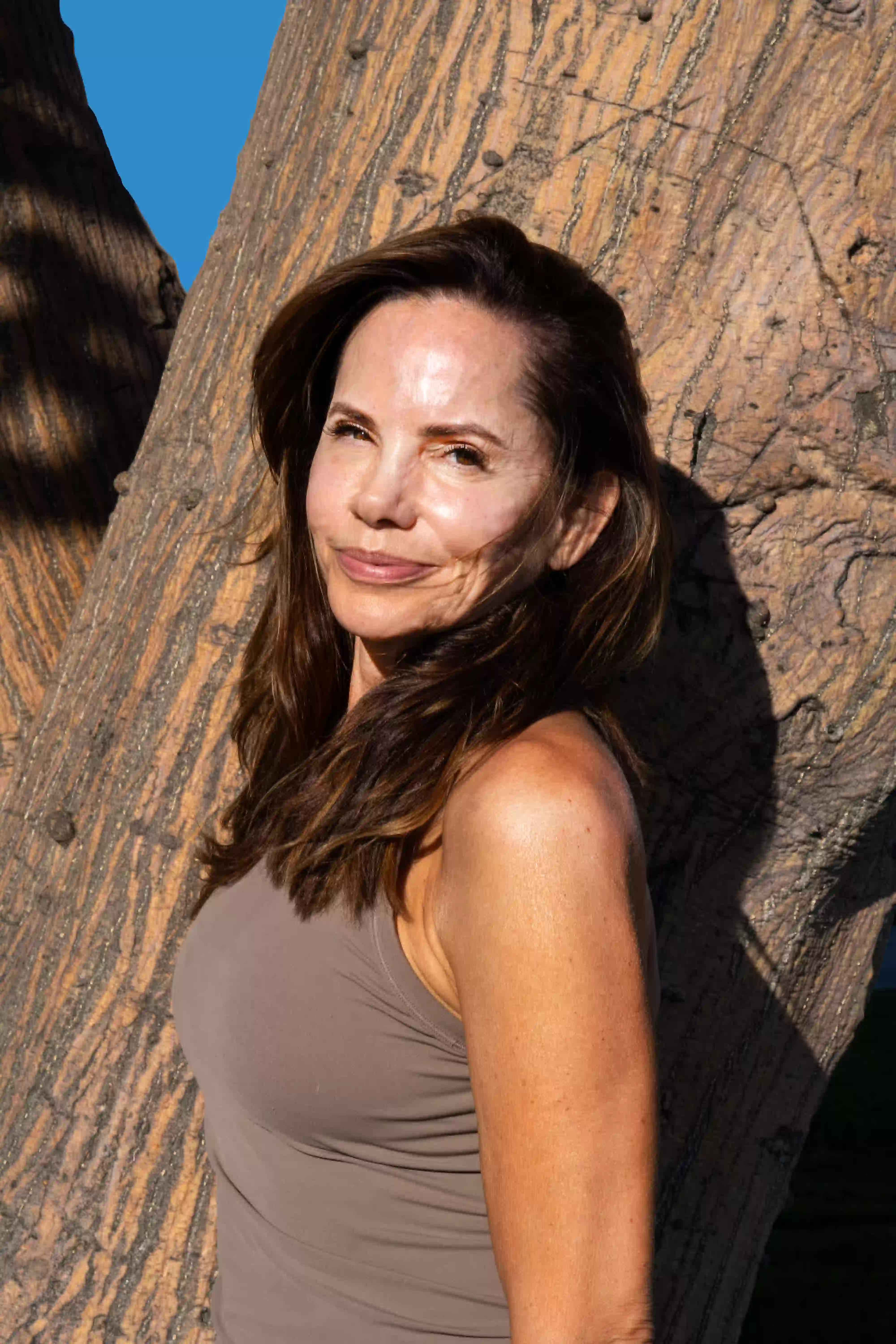 This is Cristina Clark, our founder. She is smiling, standing in front of a tree wearing a light brown tank top.