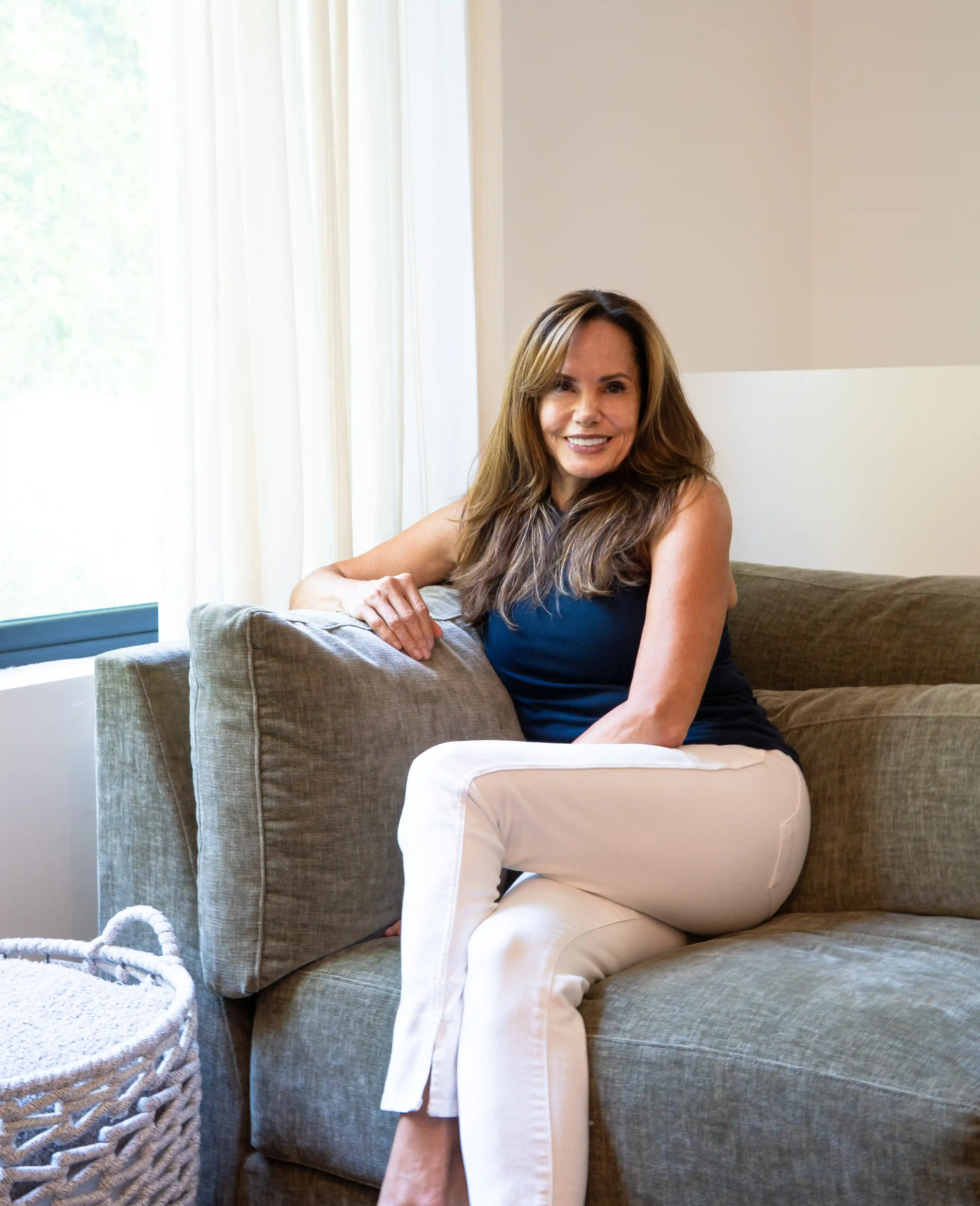 Cristina smiling confidently with long flowing hair, dressed in a navy top and white pants while sitting relaxed on a modern gray couch, radiating vitality and joy after her midlife transformation, embodying women’s empowerment, wisdom, and spiritual growth.