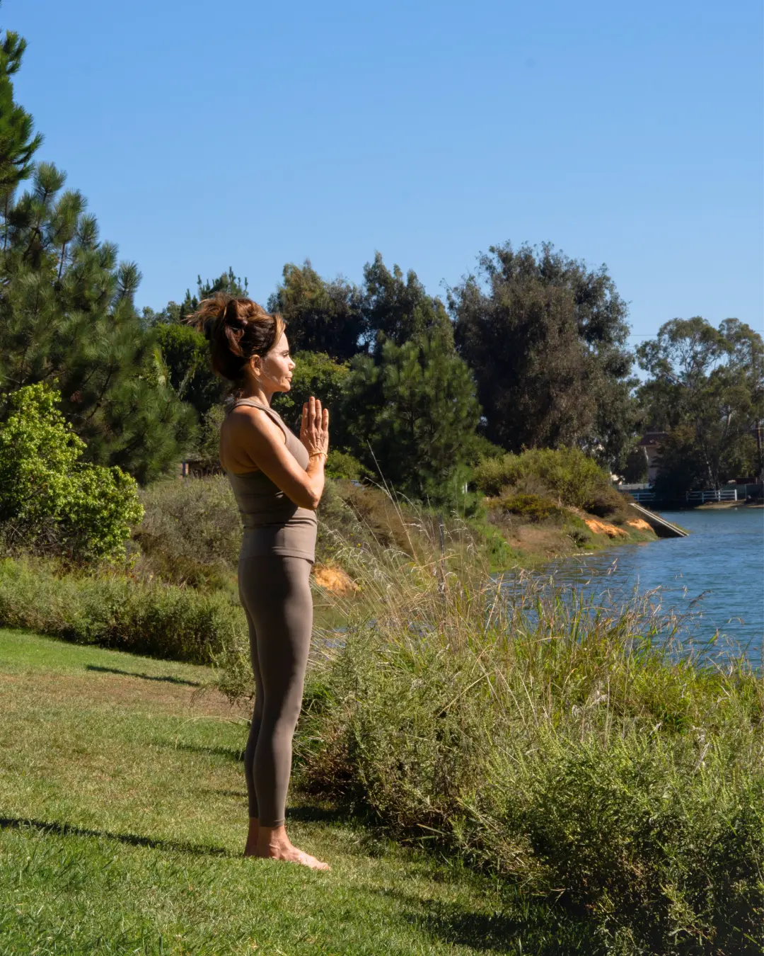 Cristina standing tall in a fitted workout outfit, hands pressed together in prayer pose by a serene lake and surrounded by trees, embracing holistic healing through yoga and meditation as she rebuilds strength, balance, and clarity during her midlife transformation.