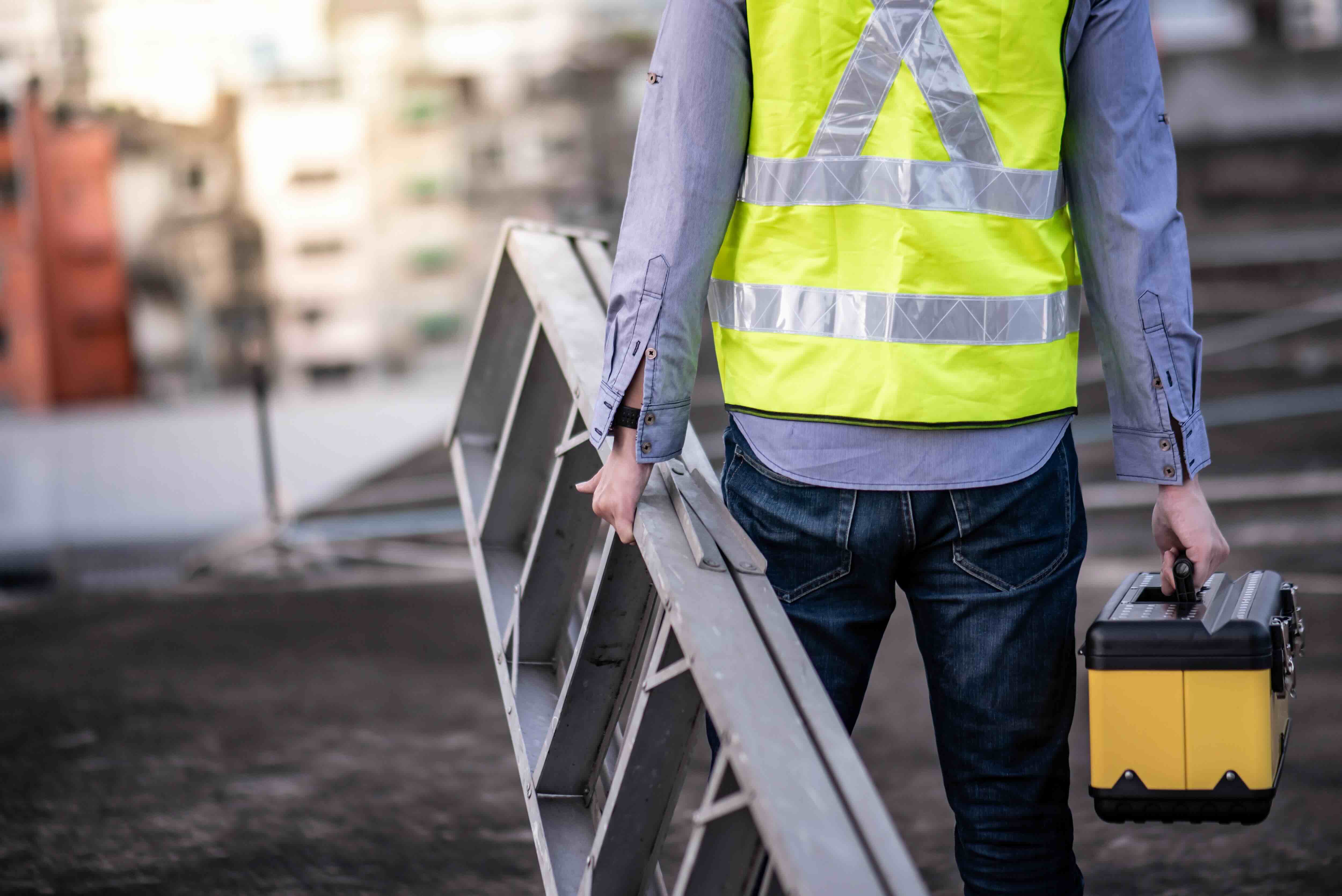 A handman holding a ladder and a toolbox wearing a reflective safety vest.