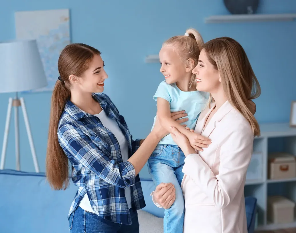 Mother handing off her young daughter to a nanny in a living room