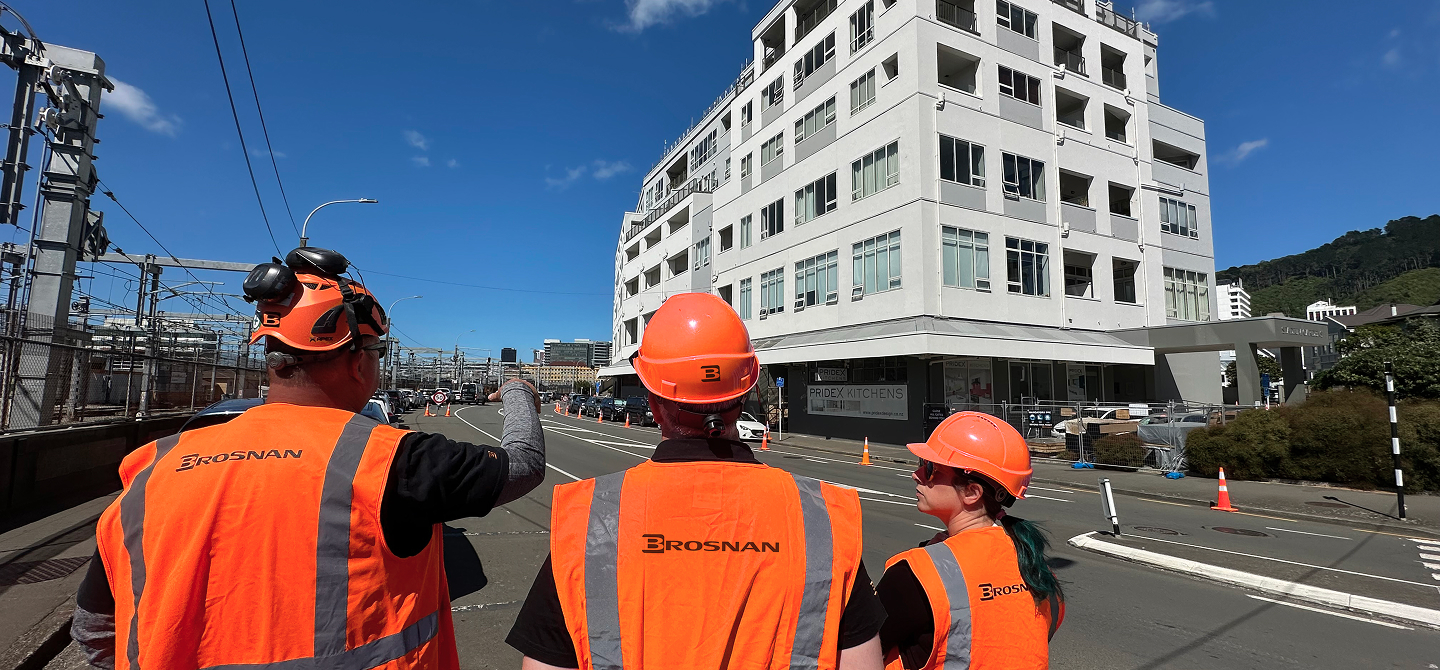 Three construction workers wearing orange safety vests and helmets stand on a street near a white building.