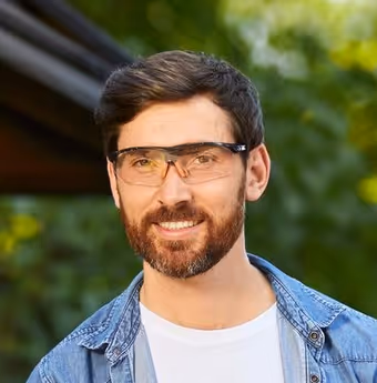 Man with dark hair and beard wearing clear safety glasses and a blue denim shirt, smiling outdoors.