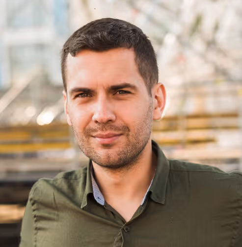 Portrait of a young man with short dark hair and stubble wearing an olive green shirt outdoors.