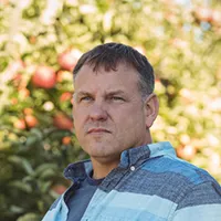 Portrait of a serious middle-aged man standing outdoors with blurry foliage and red apples in the background.