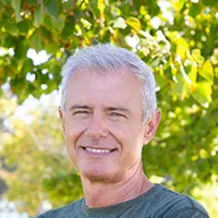 Smiling older man with gray hair standing outdoors in front of green leafy trees.