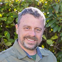 Smiling man with short gray hair and beard wearing a green shirt outdoors with green foliage background.