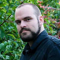 Man with a beard and shaved head wearing a black shirt, standing outdoors with green foliage in the background.