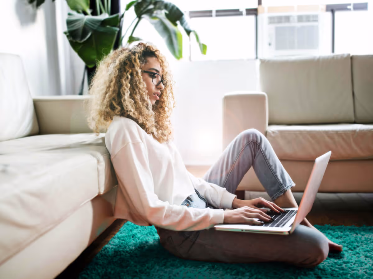 Young woman with curly hair and glasses sitting on a green rug, working on a laptop in a modern living room.