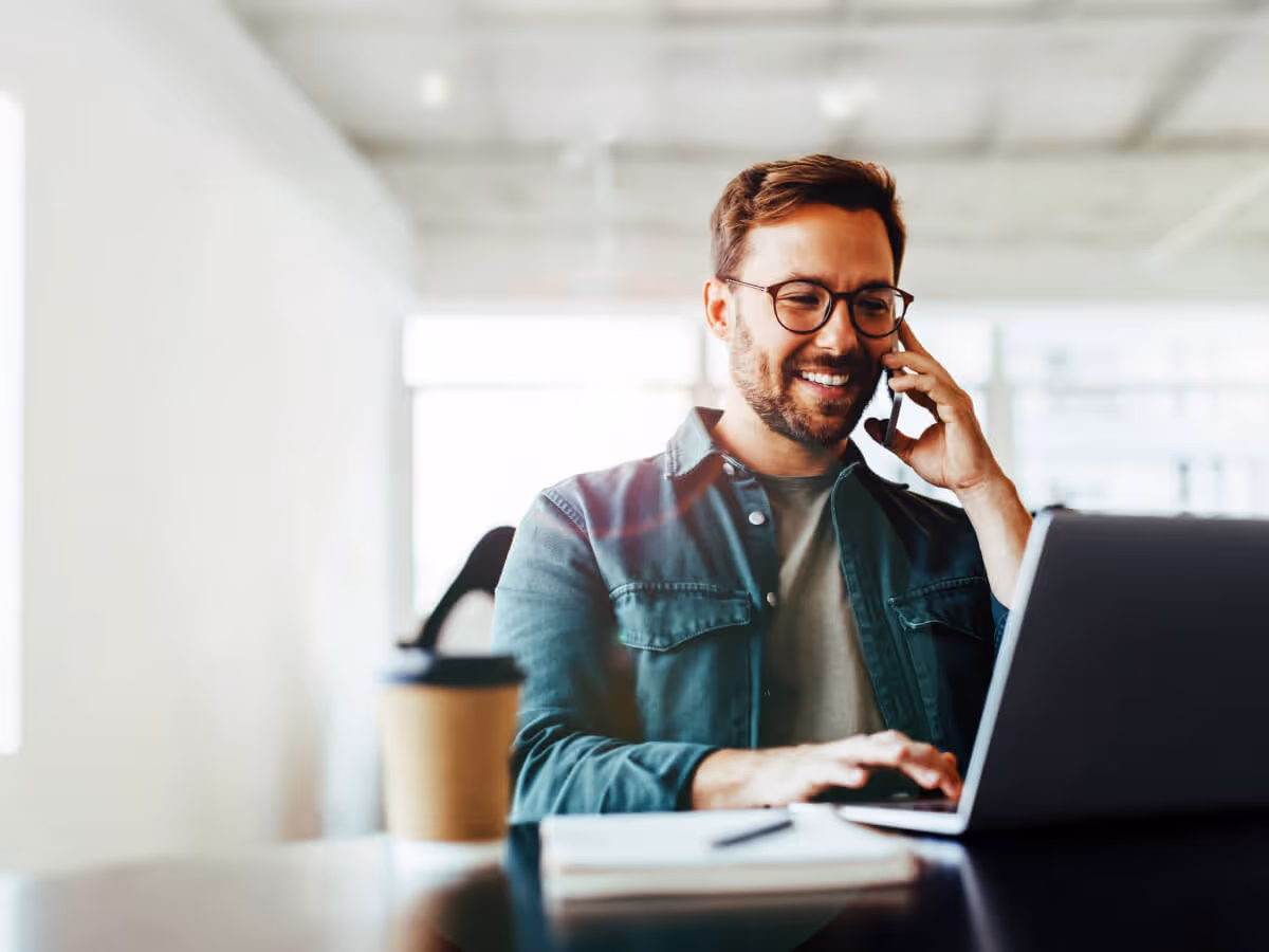 Smiling man wearing glasses talking on a smartphone while using a laptop at a desk with a coffee cup and notebook nearby.