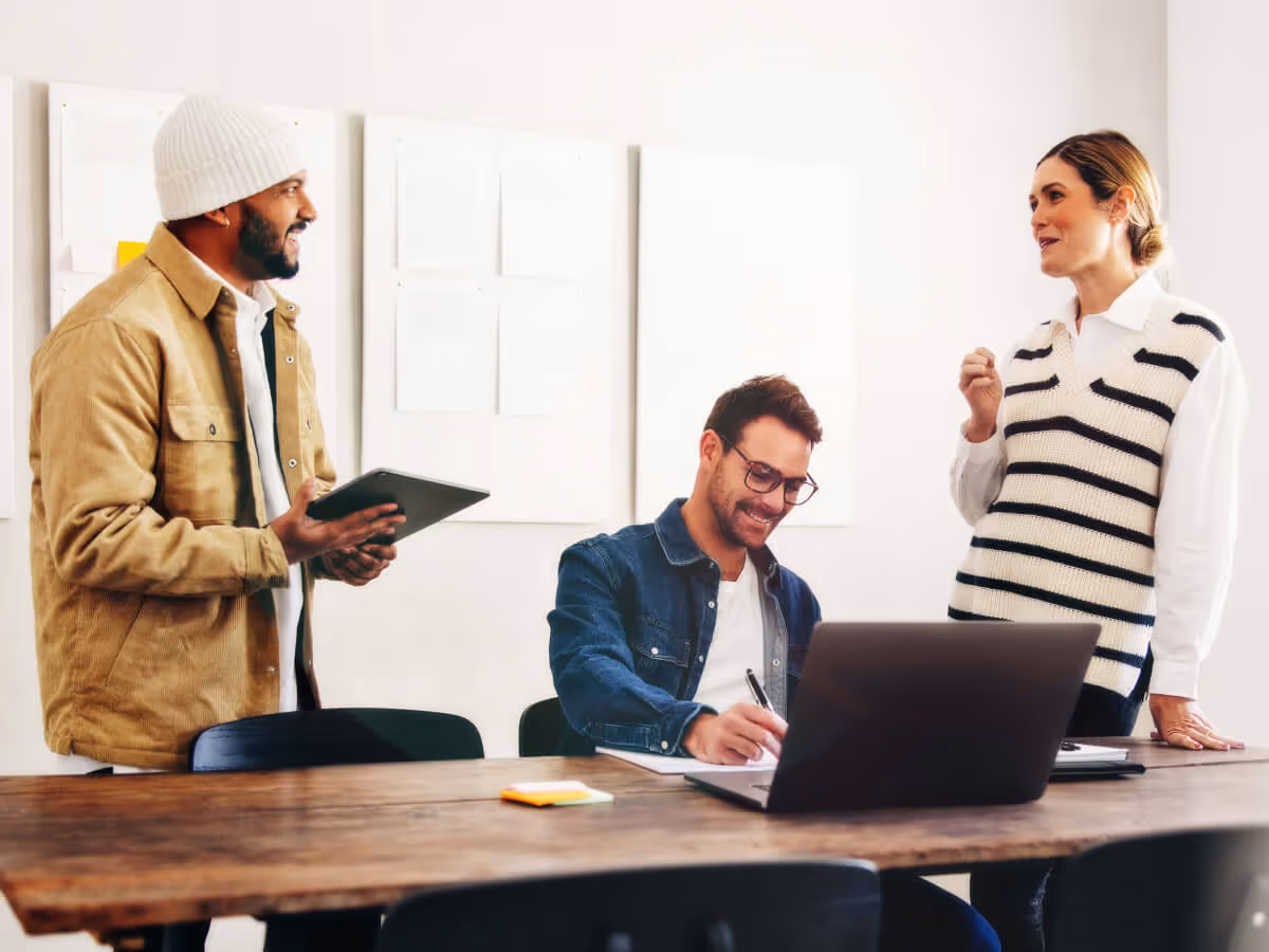 Three colleagues in a casual office setting discussing work with a laptop and tablet.
