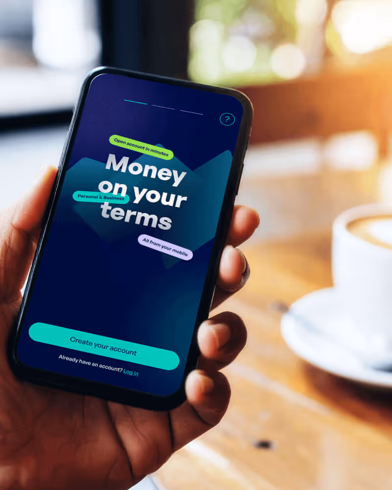 Hand holding smartphone displaying app screen that says 'Money on your terms' with options to create account or log in.