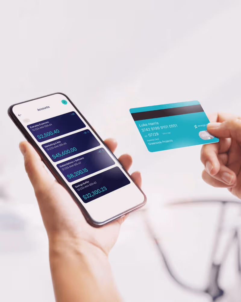Hand holding a smartphone displaying banking account balances and another hand holding a teal credit card.