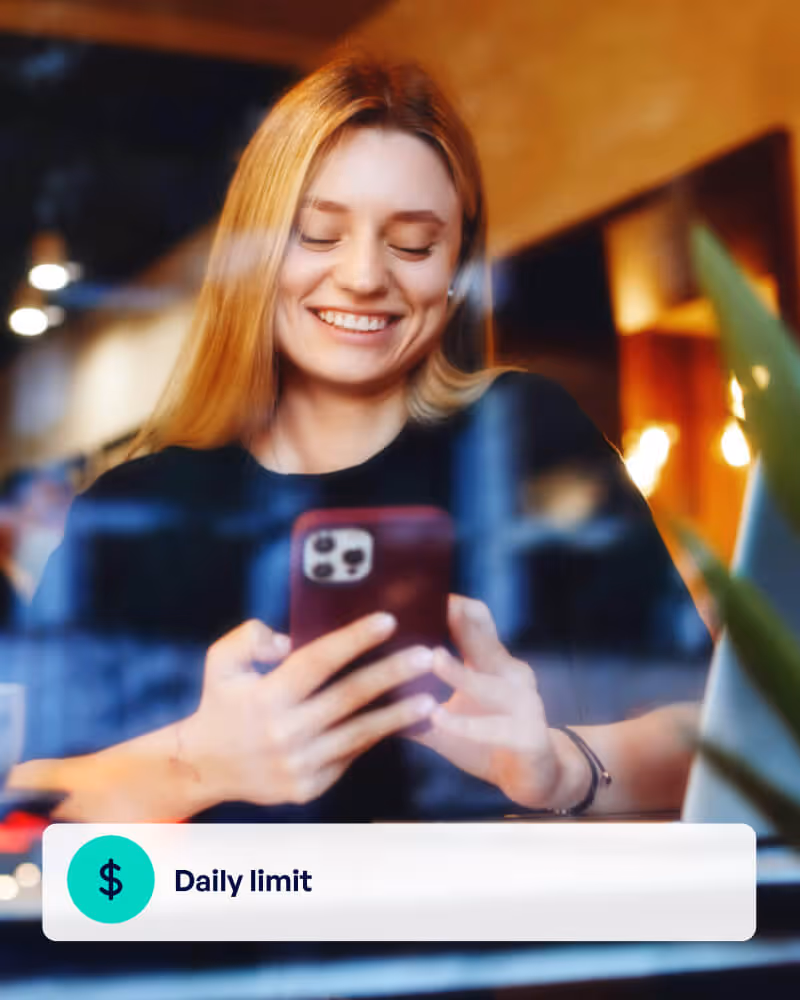 Smiling young woman looking at her smartphone screen indoors with a graphic overlay reading 'Daily limit' accompanied by a dollar sign icon.