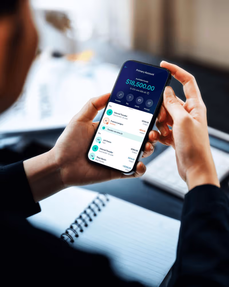 Person holding a smartphone displaying a mobile banking app with a primary account balance of $18,500 and recent transactions.