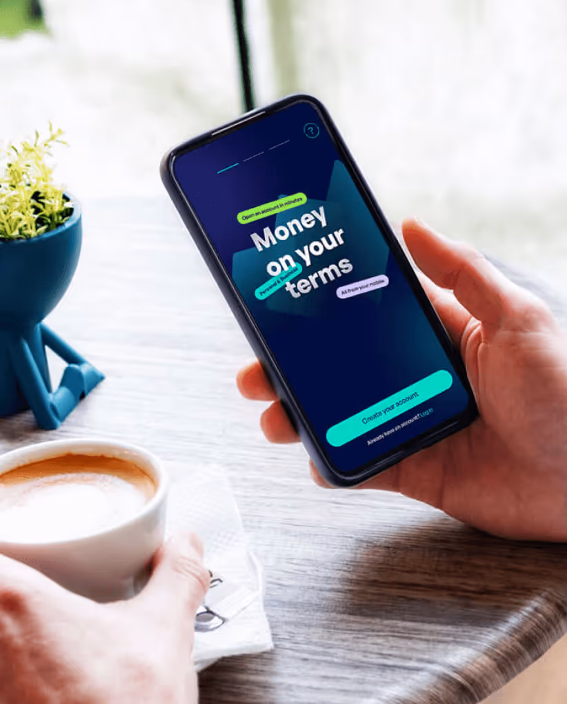 Person holding a smartphone displaying a finance app screen that reads 'Money on your terms' while sitting at a table with a cup of coffee and a small potted plant.