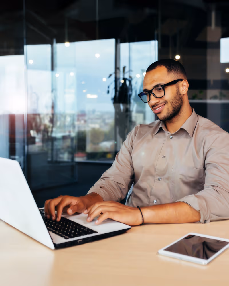 Man with glasses typing on a laptop at a desk with a tablet beside him in a modern office.