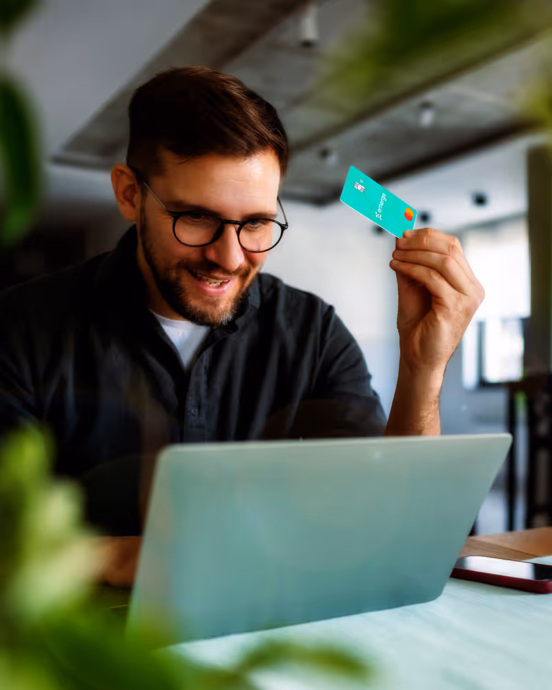 Smiling man wearing glasses holds a turquoise credit card while using a laptop at a wooden table indoors.