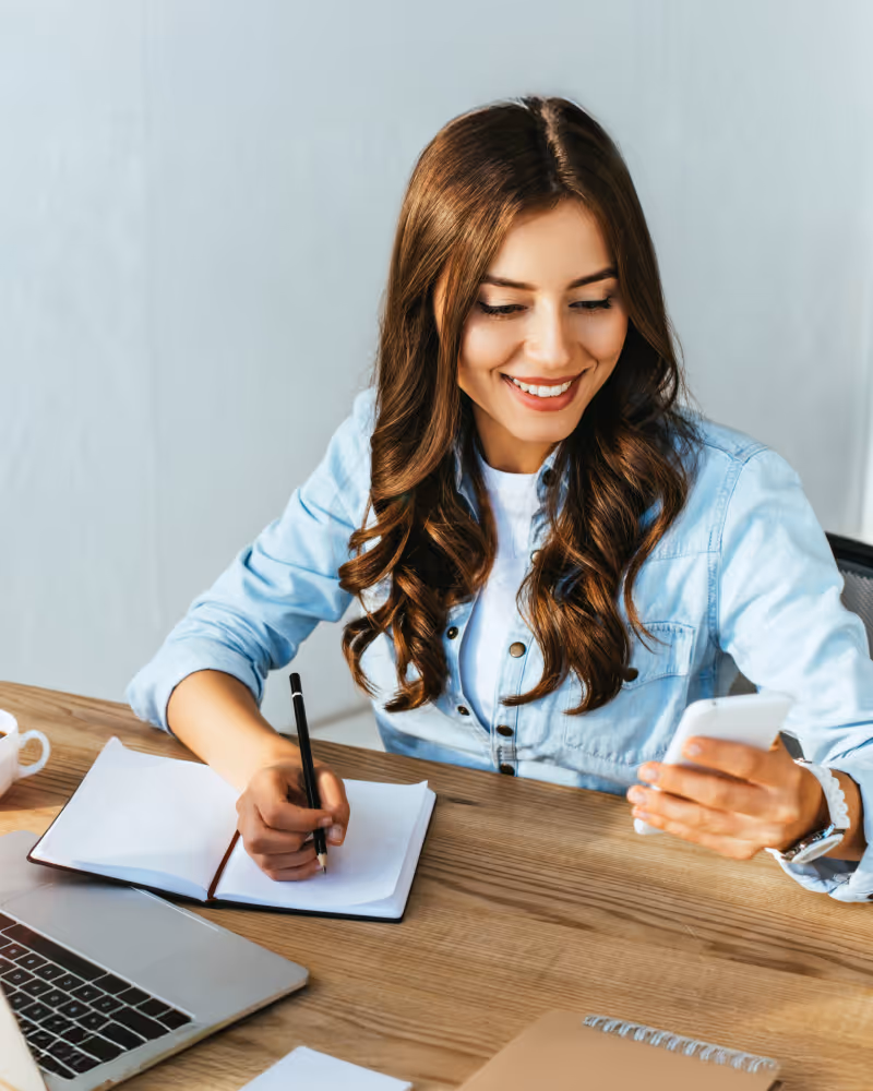 Smiling woman with brown hair in a blue shirt writing in a notebook while looking at her smartphone at a desk with a laptop and coffee cup.