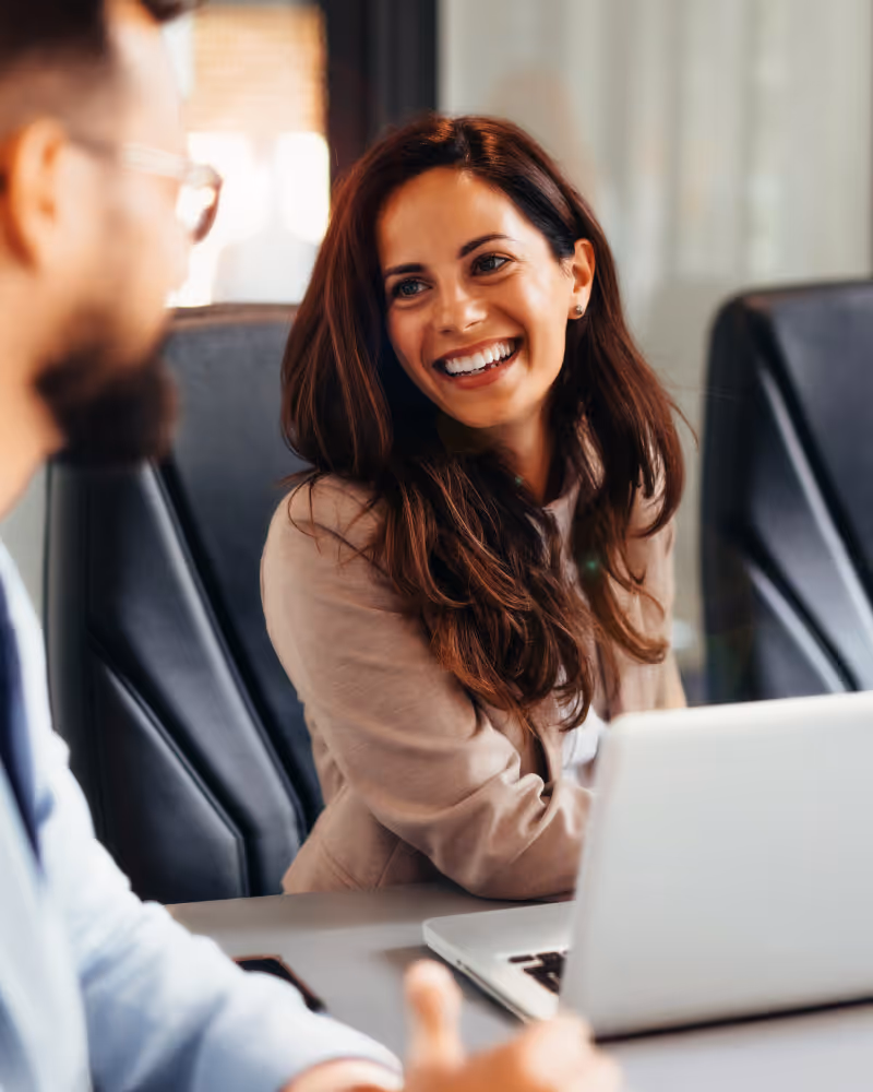 Smiling woman in business attire sitting at a table with a laptop, talking to a man wearing glasses.