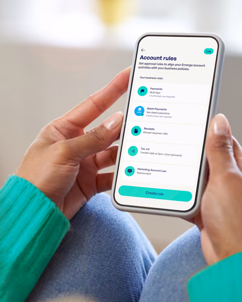 Smartphone screen displaying account rules settings with options for Payments, Batch Payments, Receipts, Tax, and Marketing Account Low, held by a person in a green sweater and jeans.