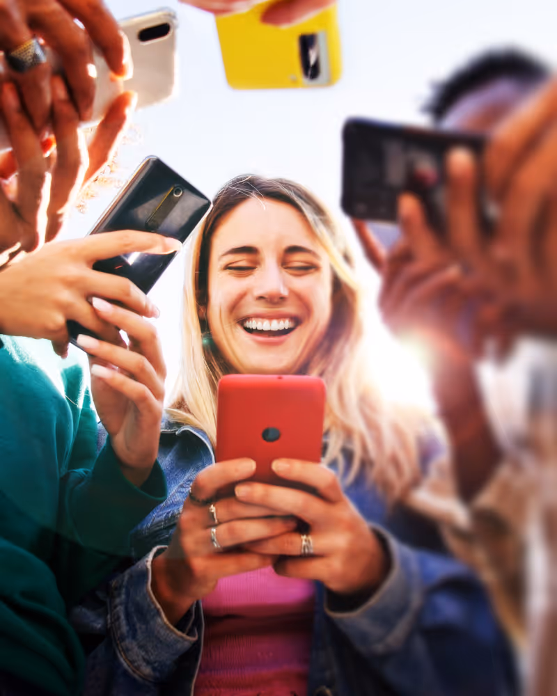 Smiling young woman in denim jacket looking at her red smartphone surrounded by others holding phones.