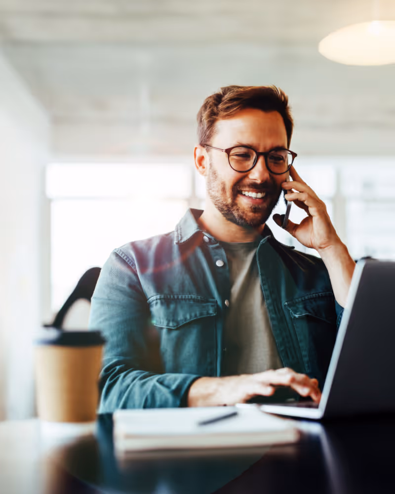 Smiling man wearing glasses talking on phone while working on laptop at desk with a coffee cup.